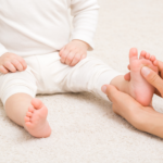 Masseur hand holding infant leg and massaging foot on carpet. Baby healthcare. Closeup. Front view.
